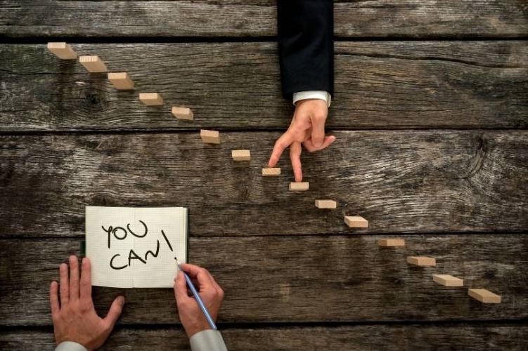A person uses their fingers to depict walking up a jenga staircase while another person holds a book open with the words "you can."