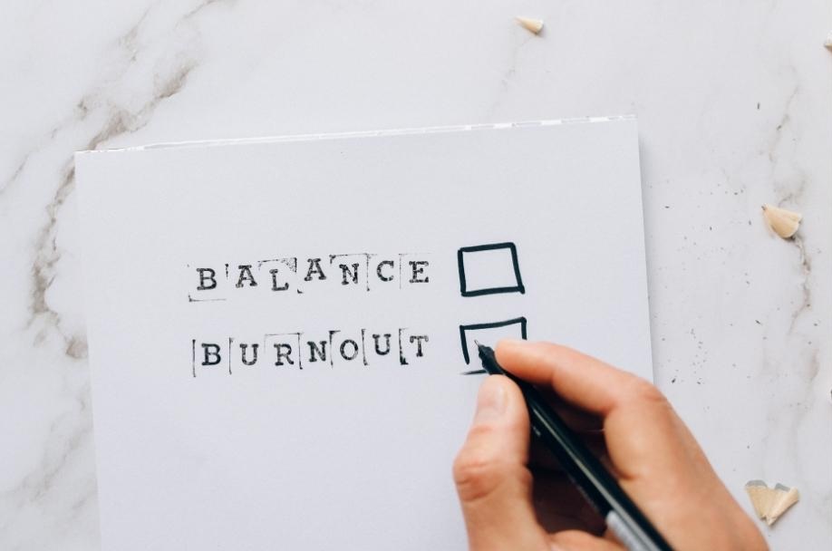 The words balance and burnout are written on a paper with a checkbox next to each word. A person is deciding which one to choose.