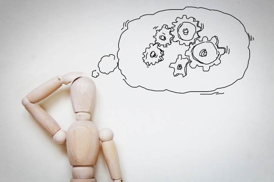 A wooden artist mannequin scratches his head beside a though bubble with turning gears in it.