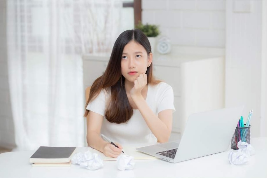 A woman sits at a desk, surrounded by crumpled papers, trying to write on behalf of her minister. 