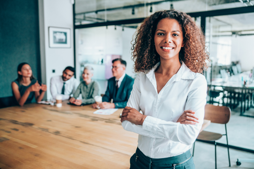 woman stands close to the camera, smiling, arms crossed. Her business team sits around a board room table behind her.