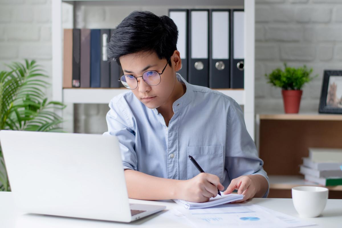 young man writing at a desk in an office, writing on a notepad and looking at a laptop screen